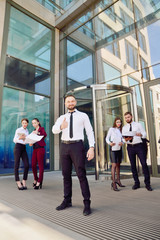 A young male businessman is enjoying a successful deal on the background of office workers. Demonstration of positive emotions and gestures. Career. Success. Shows a thumbs-up.