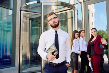 A young male manager with a tablet in his hands against the background of the girls. Office staff in the background of a multi-storey glass building. Lifestyle. Businessman