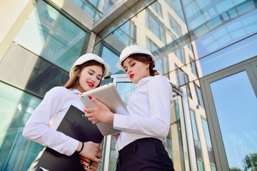 Girls in building white helmets with tablets in hands on the background of a glass office building. Women are engineers. Female professions