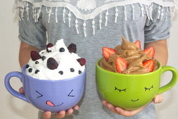 A woman holds two large mugs with drawn faces, whipped cream and berries, strawberries, sea, blackberries, blueberries. Homemade dessert.