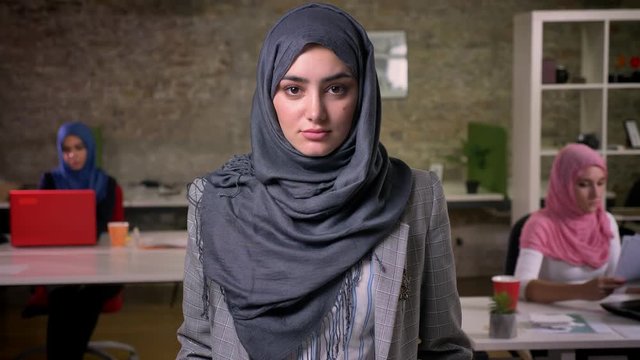 Beautiful arab woman in grey hijab is standing quietly and calmly looking at camera, brick background, working colleagues ar sitting and using theis laptops