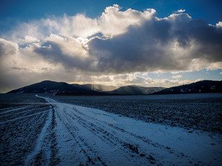 snow road in the field which is plowed leads to the mountains above which clouds and blue sky