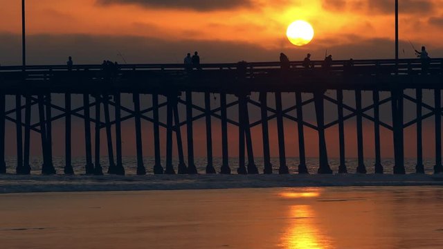 Sun in the sky above Newport Beach pier during colorful sunset while people fish from the top.