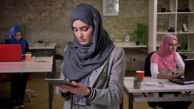 Cute arabic female in grey hijab is swwiping her gerey tablet and looking presicely at it while standing near otehr islamic working women