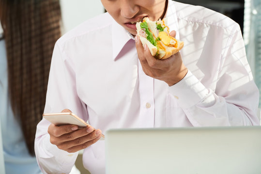 Young entrepreneur eating sandwich and reading messages on smartphone screen - Powered by Adobe