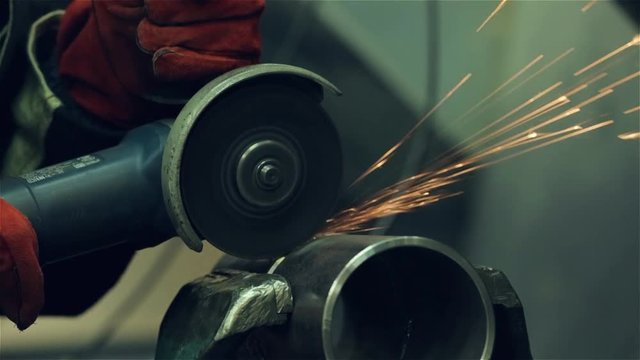 A red-gloved mechanic cleans a welded seam on a section of a steel pipe with the help of a grinding machine in the metal workshop