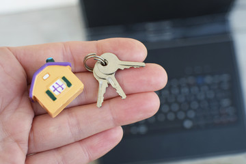 Hand holding or giving small colorful house model and keys on blurred black laptop background. Online house loan, buying or selling real estate on internet or renting new home concept.