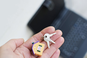 Hand holding or giving small colorful house model and keys on blurred black laptop background. Online house loan, buying or selling real estate on internet or renting new home concept.