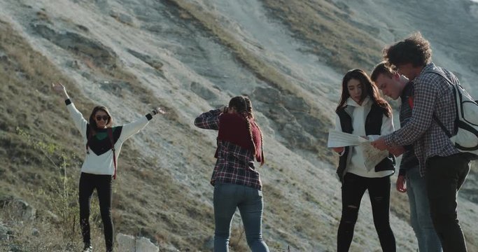 Group of multicultural friends have a trip in the middle of mountain, two girls making pictures for memories, other fiends trying to find the short way using a map. 4k