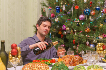 Handsome middle-aged man pours champagne into glass sitting at festive table near beautiful decorated Christmas tree in living room interior. New year holiday concept at cozy home