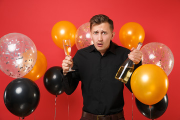 Bewildered young man in black classic shirt hold bottle, glass of champagne on red background air balloons. Valentine's, International Women's Day Happy New Year birthday mockup holiday party concept.