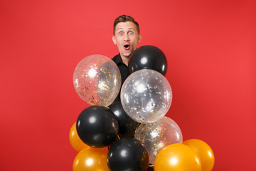 Amazed young man in black classic shirt holding air balloons, celebrating isolated on bright red background. Valentine's International Women's Day Happy New Year birthday mockup holiday party concept.