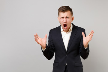 Portrait of aggressive young business man in classic black suit, shirt screaming, spreading hands isolated on grey background in studio. Achievement career wealth business concept. Mock up copy space.