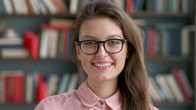 close up portrait of young pretty librarian woman smiling happy looking at camera in library bookshelf background knowledge learning.