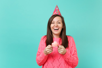 Laughing young woman in knitted pink sweater and birthday hat holding in hand cake with candle...