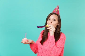 Amazed young woman in knitted pink sweater and birthday hat with playing pipe holding in hand cake...