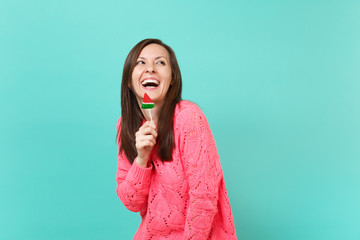 Fototapeta premium Laughing cheery young woman in knitted pink sweater looking up, holding watermelon lollipop isolated on blue turquoise wall background, studio portrait. People lifestyle concept. Mock up copy space.