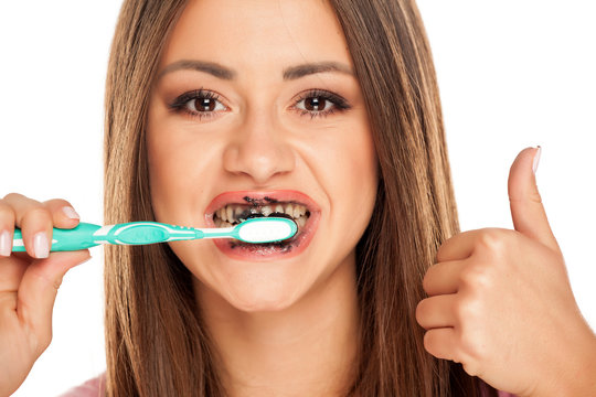 young woman brushing her teeth with black active charcoal toothpaste on white background and showing thumbs up