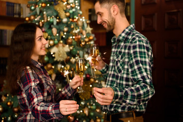 Merry Christmas and Happy New Year! Attractive young couple is celebrating holiday at home together, drinking champagne and smiling with Bengal lights in hand