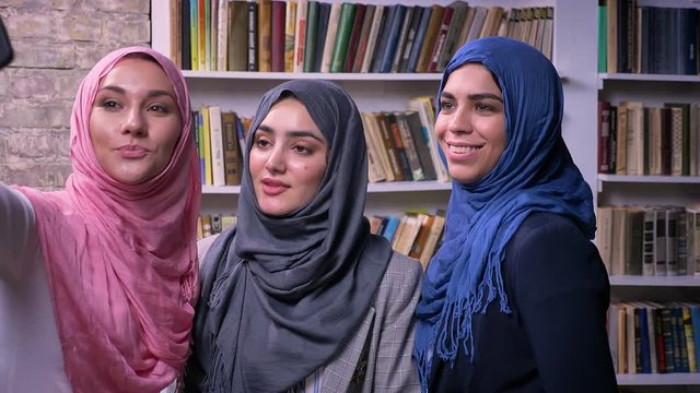 Three pretty hijab muslim females are taking pictures and selfie with phone while standing indoor, books on background, happy mood