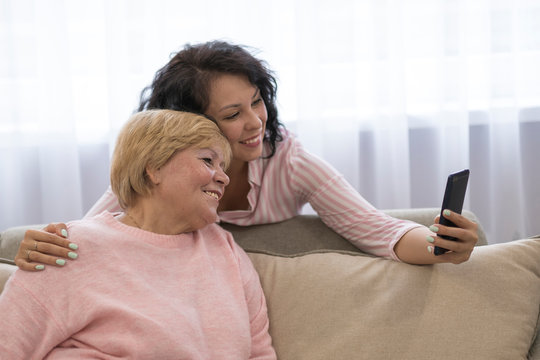 Portrait Of Old Mother And Mature Daughter Hugging At Home. Happy Senior Mother And Adult Daughter Embracing With Love On Sofa. Cheerful Woman Hugging From Behind Older Mom And Looking At Each Other.