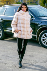 Beautiful brunette in a light-colored fur coat and black pants walks down the street in front of the car on an autumn sunny day