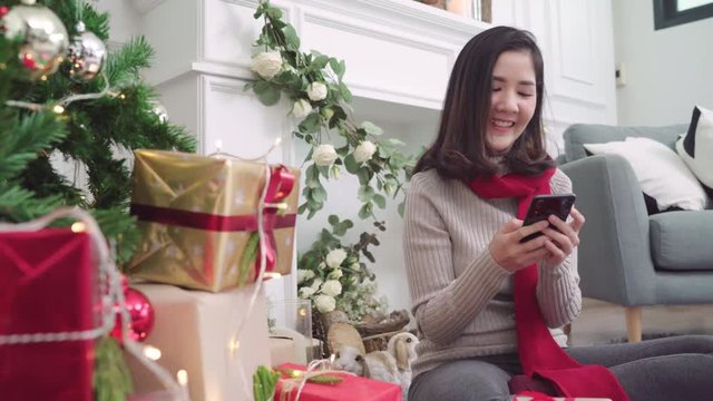 Cheerful happy young Asian woman using smartphone to check social media in her living room at home in Christmas Festival. Lifestyle women celebrate Christmas and New year concept.