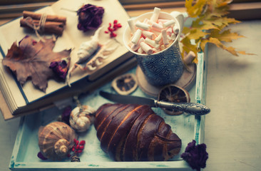 a cup of coffee with marshmallows,crroissant,books,seashells,dried roses and autumn leaves/coffee on the  windowsill with autumn view/cozy autumn