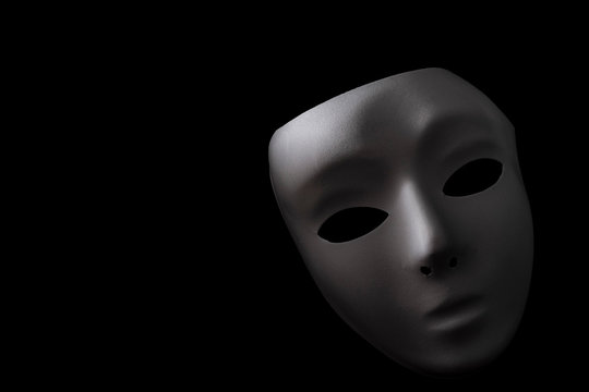 Shadowy disguise, secret society and creepy emotionless figure concept with a vintage white blank mask with dramatic light and dark shadow isolated on black background with copy space