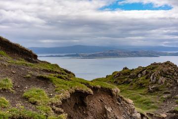 Erdig felsige zum Teil mit Gras bewachsene Berglandschaft mit Blick auf das ruhige Meer und einer Landzunge darüber blauer Himmel mit Wolkendecke