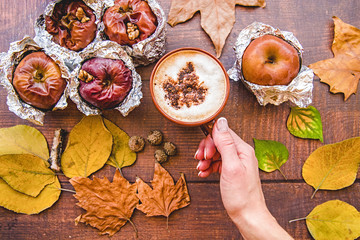 hand with a cup of coffee and backed apples in foil with autumn leaves on wooden background