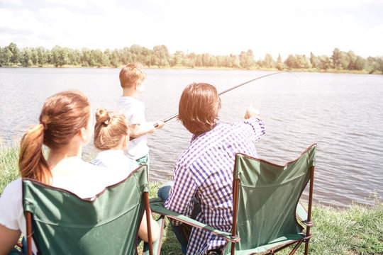 A picture from the back. Small boy is holding a fish-rod and trying to catch some fish in the river. His dad is guiding him and pointing forward. Woman is holding small girl on her lap.