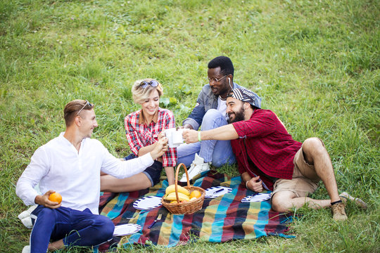 Group of young cheerful coworkers using lunch break time having fun outdoors on green lown, celebrating the start of a new project with fruit drinks