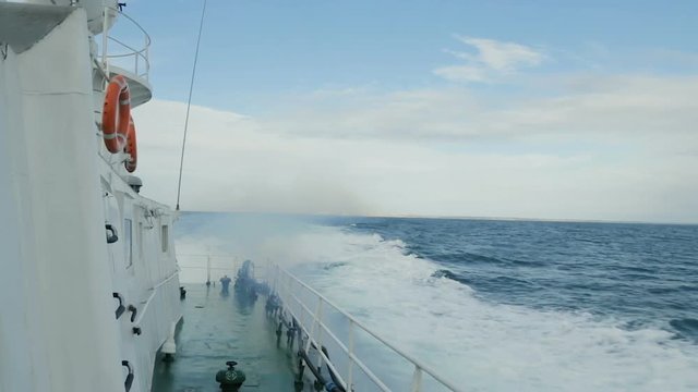View from deck of a ship as the waves are splashing. Military ship is swimming at sea. Boat is Floating on the Waves and Leaves a Trail in Sea. Red life buoys on the stern of the ship.
