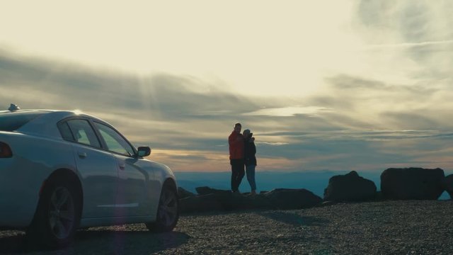Caucasian couple standing on a cliff, taking selfies against mountain background, car in a foreground. 4K UHD