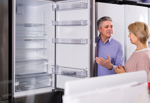 joyous mature married couple in shop of household appliances are choosing home modern refrigerator