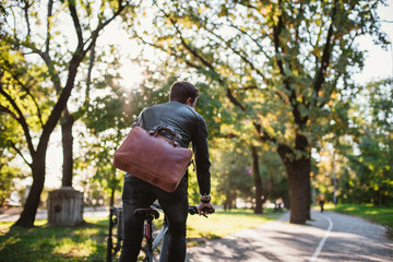 Young businessman with leather bag riding bicycle to city park