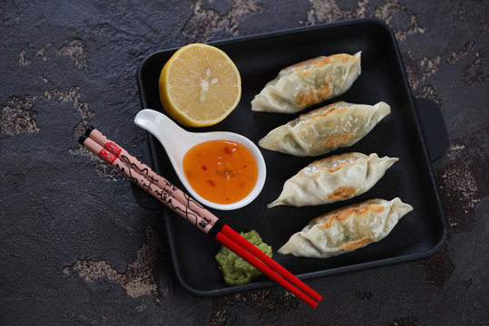 Cast-iron serving pan with fried korean dumplings, dipping sauces and eating sticks. High angle view over brown stone background
