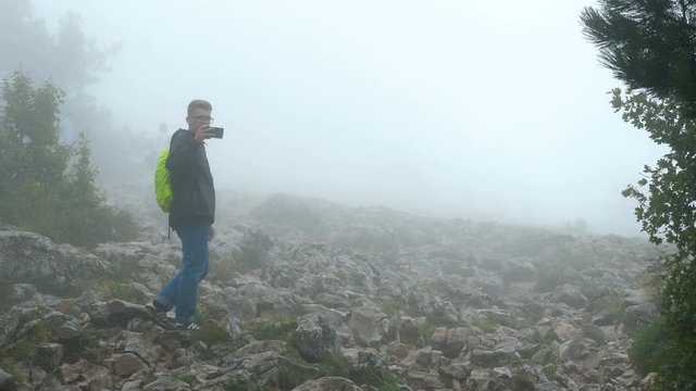 A group of tourists is walking along a gloomy foggy mountain covered with forest. A young man behind the group and taking a photo on a mobile phone.