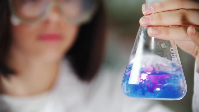 Young woman in chemical laboratory holding a flask liquid and shaking it. The liquid changes color. Focus on the flask.