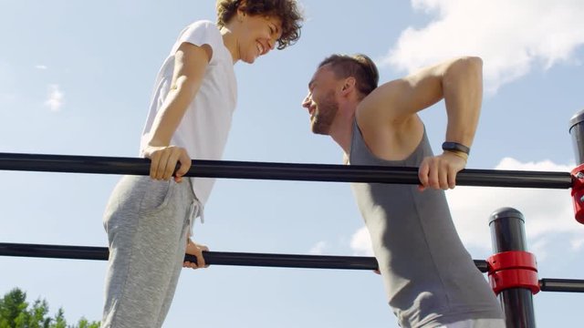 Low angle view of happy young couple in sportswear smiling and doing dips on parallel bars outdoor on sunny summer day