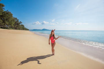 Happy young woman with long hair in red swimsuit wearing funny christmas reindeer antlers holding something imaginary standing on the beach by the sea with blue water and sky on Phuket island,Thailand