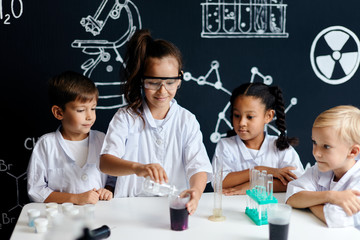 Multiethnic group of 7 years old kids in white lab coats doing a chemical experiment in laboratory...