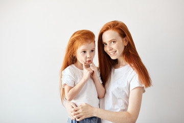 Close-up portrait of smiling mother hugging cute four year old daughter isolated on white background. Two red headed similar females of different generations with red hair down looking at camera.