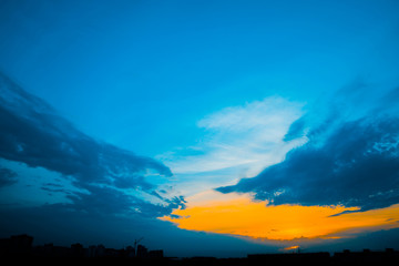 Atmospheric blue cloudy sky behind silhouettes of city buildings. Cobalt and orange background of sunrise with dense clouds and bright yellow sunny light for copy space. Cyan heaven above clouds.