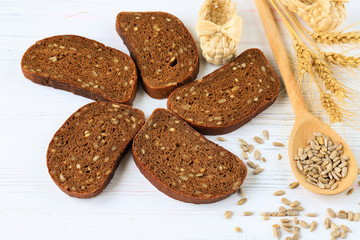 Rural still life on a white wooden background - sliced rye brown bread, sunflower ,seeds on a light wooden spoon and ears of wheat