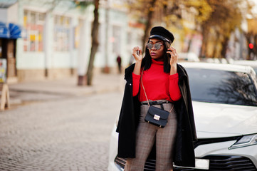 African american fashion girl in coat, newsboy cap and sunglasses posed at street against white business car.