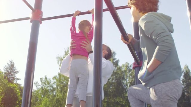 Dad in sportswear lifting little daughter on arms and helping her to do pull ups on bar while mother smiling and looking them at outdoor jungle gym on sunny day