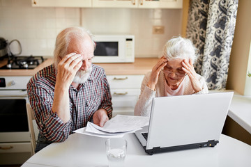 Worried depressed mature couple sitting at dining table at home, aged retired man and woman sitting...
