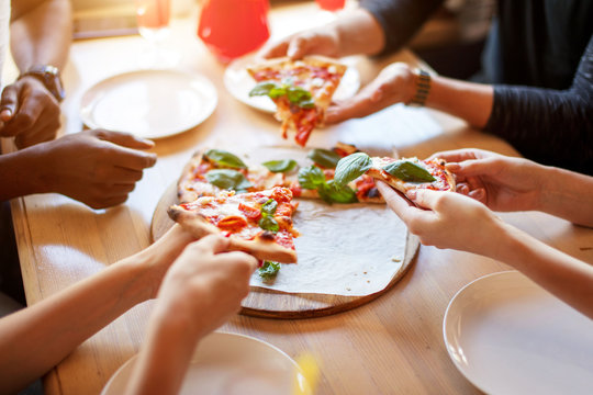 Cheerful multiracial friends eating delicious vegetarian organic pizza with basil leaves at pizza hut , close up hands .